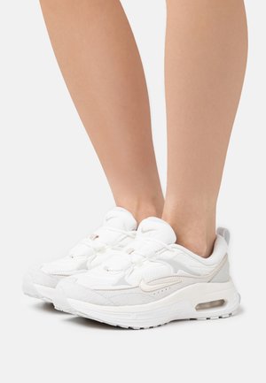 Scarpe sportive bianche con una combinazione di materiali lisci e testurizzati. I dettagli notevoli includono accenti grigio chiaro e un'unità di ammortizzazione ad aria.