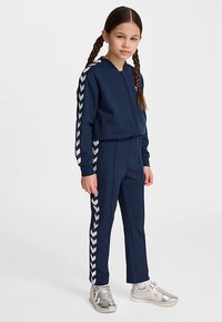 Marineblauw trainingspak met een jas met ritssluiting en bijpassende broek. De jas heeft witte chevronstrepen langs de mouwen; gecombineerd met metalen zilveren sneakers.