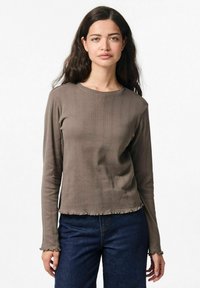 Maglia a maniche lunghe marrone a coste con scollo rotondo e orlo sfrangiato, caratterizzata da righe verticali testurizzate e polsini aderenti. Abbinata a jeans scuri.