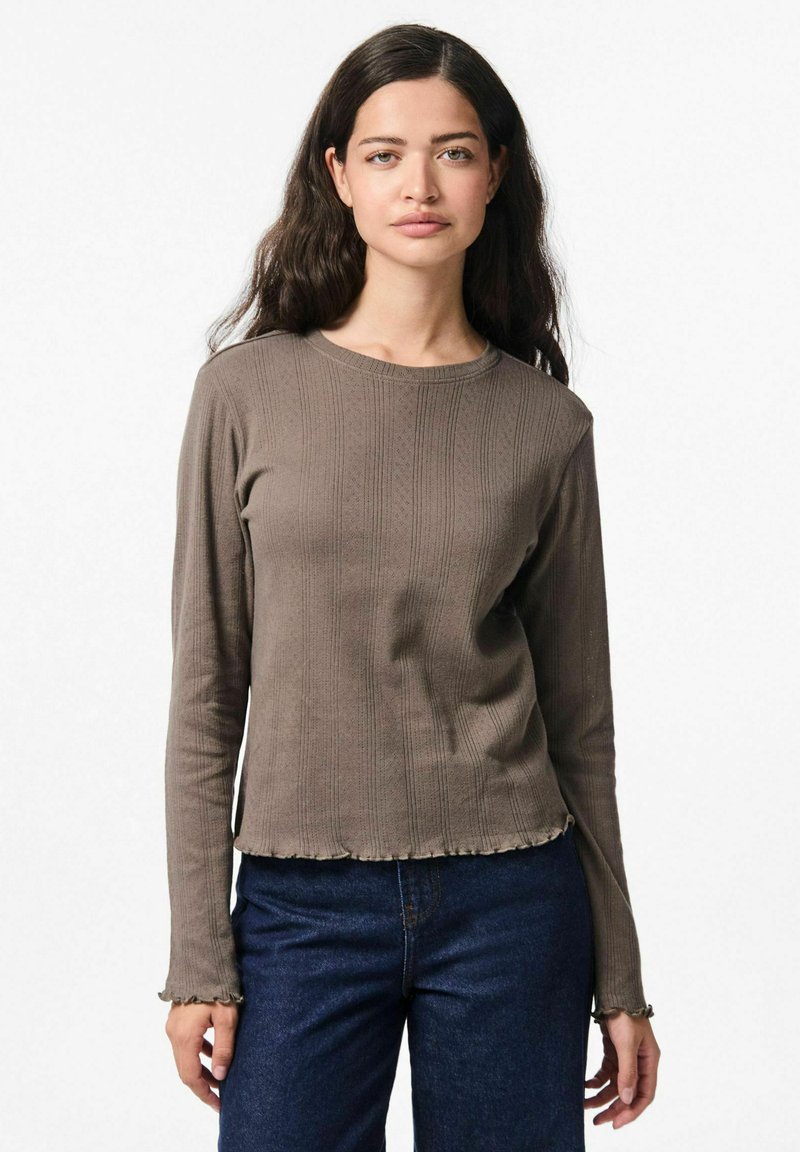 Maglia a maniche lunghe marrone a coste con scollo rotondo e orlo sfrangiato, caratterizzata da righe verticali testurizzate e polsini aderenti. Abbinata a jeans scuri.