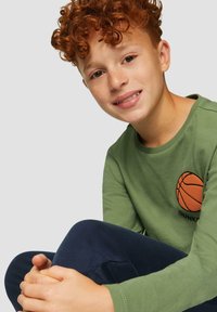 Groene longsleeve T-shirt met een basketbalafbeelding en de tekst "DUNKING," gecombineerd met donkerblauwe jeans. Kruidige haren en een neutrale achtergrond.