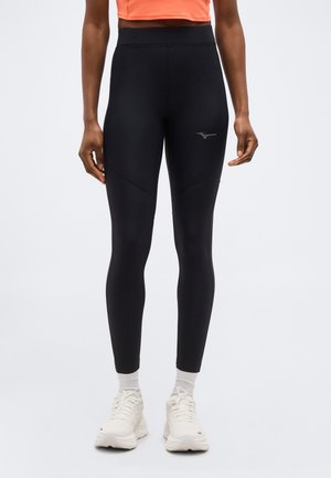 Άτομο που φοράει μαύρο κολάν ψηλόμεσο με λογότυπο στο μηρό, λευκές κάλτσες, λευκά αθλητικά παπούτσια και πορτοκαλί κοντό τοπ, στέκεται μπροστά σε απλό φόντο.