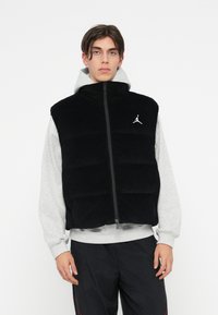 Čierny pížmo vest s vertikálnymi prešívaniami, vysokým golierom a strieborným logom Jordan na ľavej hrudi, nosený cez šedú mikinu.