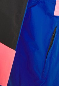 Bunda s barevnými bloky v černé, růžové a modré barvě. Hladká textura s kapsou na zip v modré části.