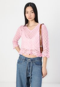 Rožinis megztas trumpas megztinis su V formos iškirpte, puoštas dekoratyviniu nėriniuotu raštu. Derinamas su aukštos liemens mėlynais džinsais ir rudu peties dirželiu.