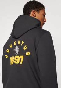 Svart huvtröja med ett grafiskt tryck som visar "JUVENTUS" och "1897" i gult, samt en grå hästsymbol på baksidan. Texturerad tyg.
