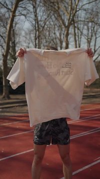 Άτομο κρατάει ανοιχτόχρωμο T-shirt με την επιγραφή «No Frills, No Hype, Start Running» σε αθλητικό στίβο με γυμνά δέντρα στο φόντο.