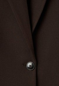 Dettaglio di un blazer marrone con rever a punta, tessuto testurizzato e un pulsante nero lucido con design a quattro fori.