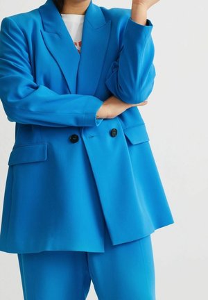 Personne portant un blazer bleu vif à double boutonnage et un pantalon assorti, les bras croisés, sur un fond clair uni.