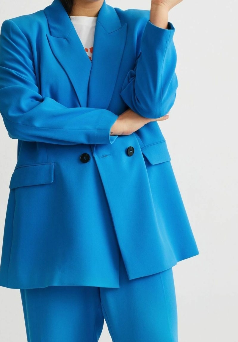 Personne portant un blazer bleu vif à double boutonnage et un pantalon assorti, les bras croisés, sur un fond clair uni.