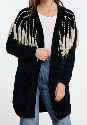 Woman wearing black knit cardigan with white fringe details on shoulders, white shirt, and blue jeans, holding cardigan front.