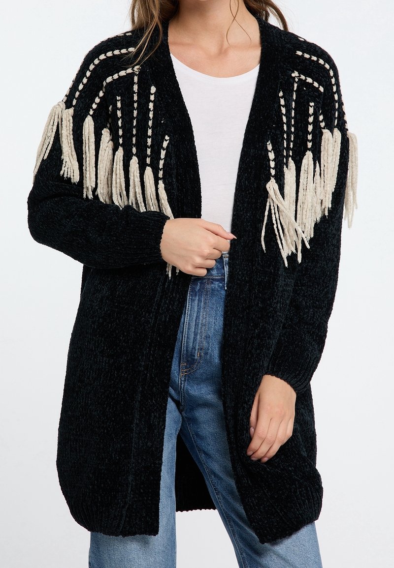 Femme portant un cardigan en maille noir avec des détails de franges blanches aux épaules, une chemise blanche et un jean bleu, tenant le devant du cardigan.