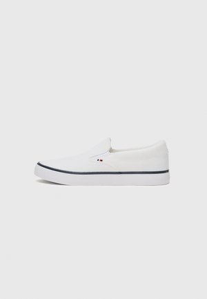 Zapatillas blancas sin cordones, hechas de tejido texturizado, con una suela plana blanca y una franja oscura azul contrastante. Presentan un pequeño logo en el lateral.