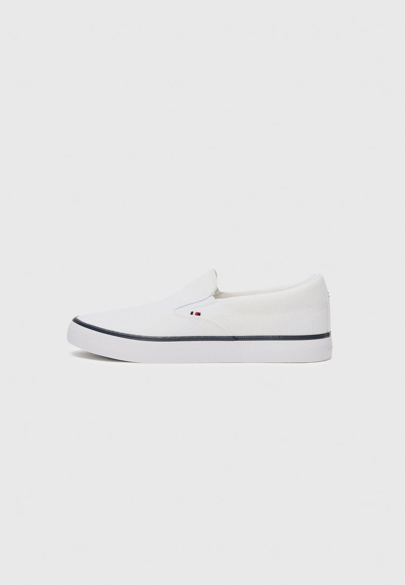 White slip-on shoes made of textured fabric, with a flat white sole and a contrasting dark blue stripe. Features a small logo on the side.