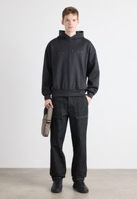 Felpa grigia con logo "Targa", pantaloni in denim neri, sneakers nere, mentre tiene una borsa grigia. Linee pulite e design casual. Texture semplici.