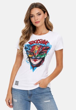 Frau mit langen, welligen Haaren, trägt ein weißes T-Shirt mit einem bunten Katzenkopf und dem Schriftzug "ROCK SLAVE", kombiniert mit zerrissenen blauen Jeans.