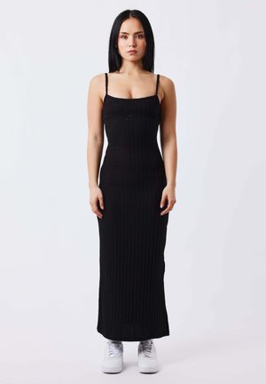 Mujer con cabello largo y negro que lleva un vestido maxi negro sin mangas de canalé y zapatillas blancas, posando sobre un fondo claro y liso.
