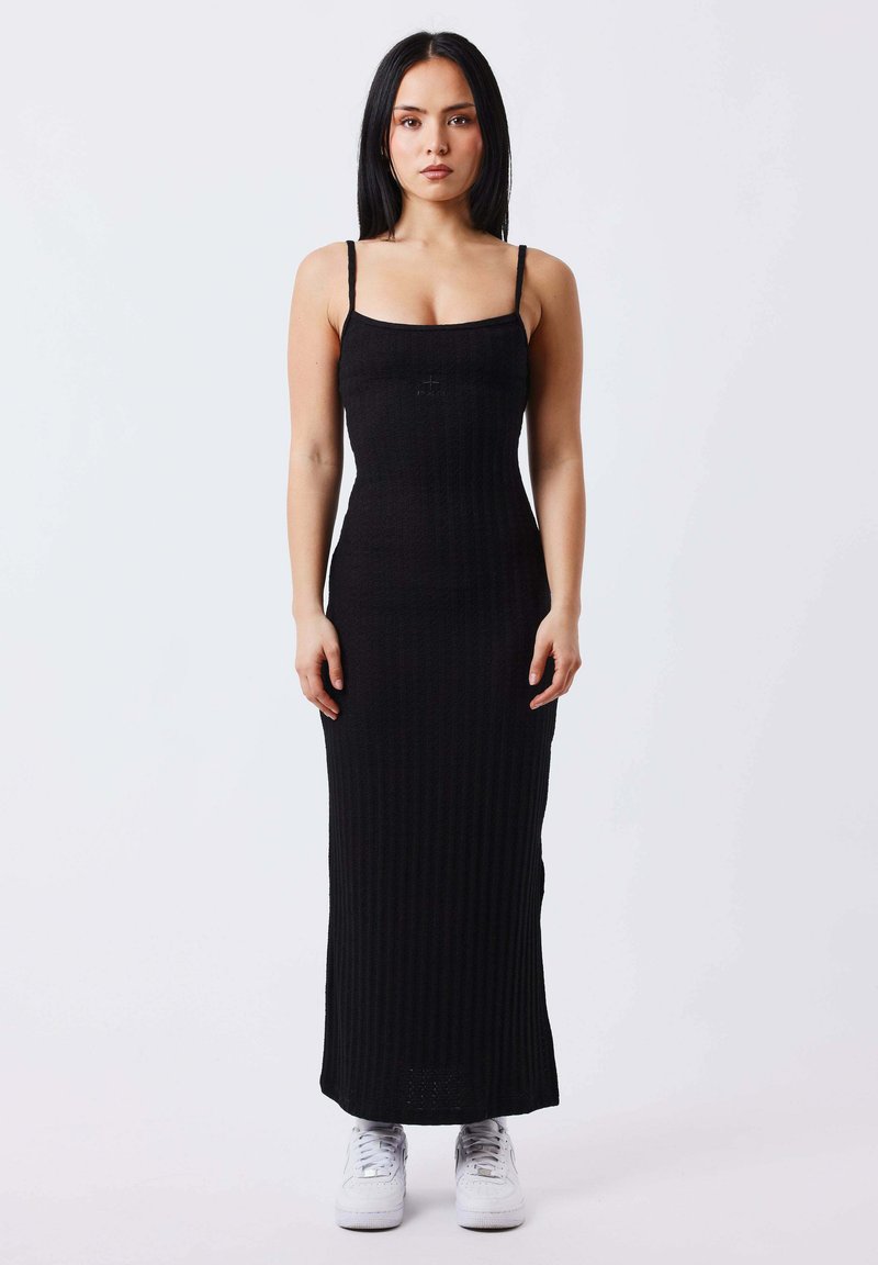 Femme aux longs cheveux noirs portant une robe maxi noire côtelée sans manches et des baskets blanches, debout sur un fond uni clair.