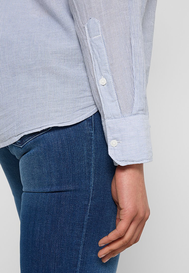 Blauw-witte gestreepte blouse met lange mouwen en knoopmanchetten, gemaakt van lichte stof, gecombineerd met aangesloten blauwe spijkerbroek.