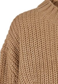 Gros plan d'un pull épais en maille couleur chameau montrant un motif côtelé et un col roulé en tissu tricoté.