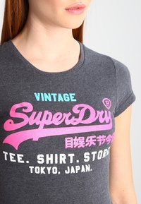 Grått slips i t-shirt med korta ärmar. Framsidan har "SuperDry"-logotypen i rosa och blått, med vit text. Tillverkad av mjukt bomullsmaterial.