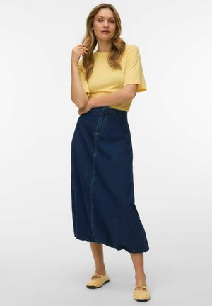 Woman wearing a yellow short-sleeve top, high-waisted dark blue denim midi skirt, and pale yellow ballet flats standing against white background.
