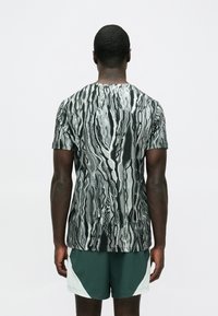 Koszulka z krótkim rękawem w czerni i bieli z abstrakcyjnym wzorem spiralnym, okrągłym dekoltem i miękkim wykończeniem materiału. Noszona z zielonymi szortami.