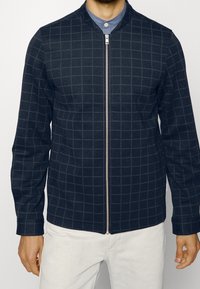 Uomo che indossa una giacca blu navy con zip e un motivo a griglia grigio chiaro sopra una camicia azzurra con bottoni e pantaloni bianchi, con le mani rilassate ai fianchi.