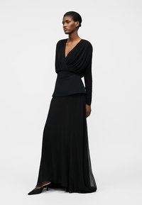 Wysoka kobieta ubrana w długą czarną sukienkę z drapowanym dekoltem w serek, dopasowaną talią i czarnymi szpiczastymi szpilkami, stojąca na jednolitym tle.