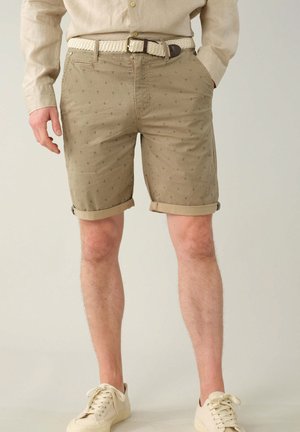 Shorts kaki avec revers roulés, présentant un motif subtil de petites ancres sombres. Portés avec une ceinture tressée crème et des baskets claires.