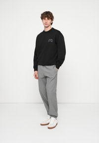 Sudadera negra con cuello redondo y logo, combinada con pantalones plisados gris claro y zapatillas blancas con detalles marrones. Diseño minimalista.