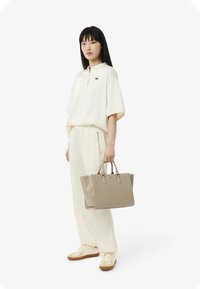 Maglietta a girocollo beige chiaro e pantaloni abbinati, accompagnati da una borsa a tracolla leggera marrone con trama. Le scarpe da ginnastica sono di colore crema e nocciola.