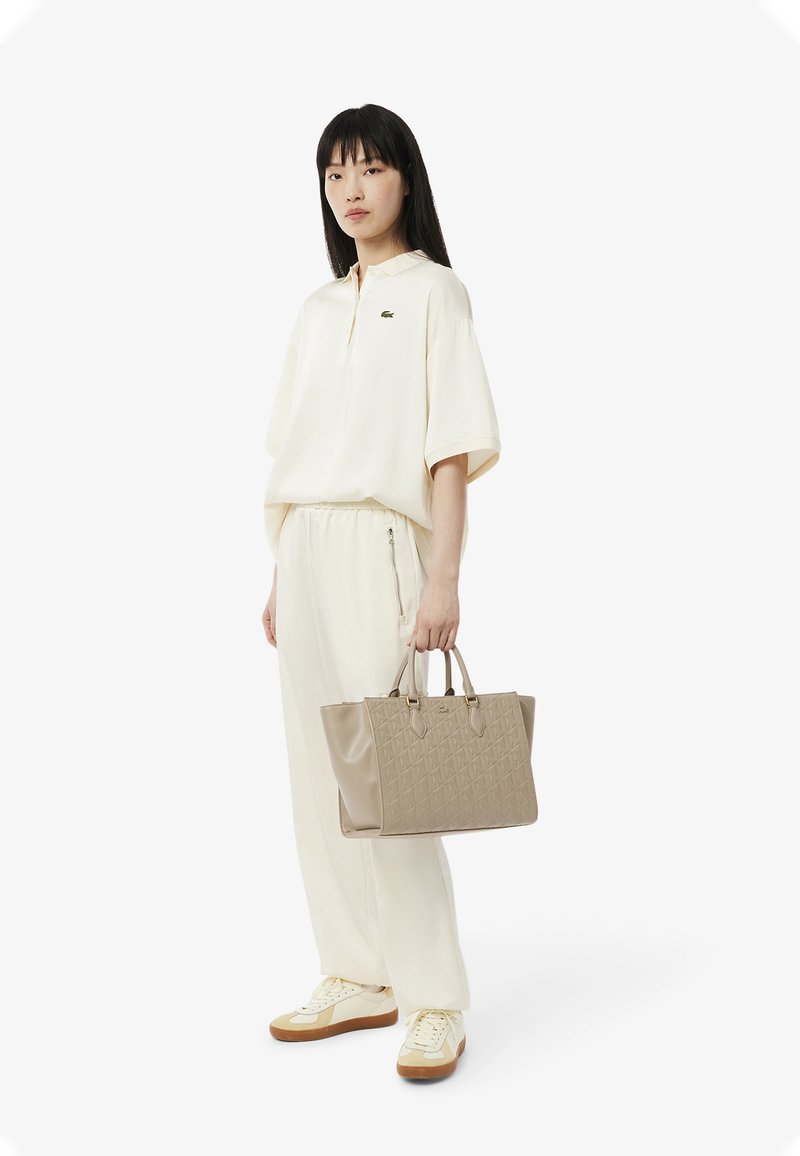Licht beige crewneck shirt en bijpassende broek, gecombineerd met een gestructureerde lichtbruine handtas. Sneakers zijn crème en tan.