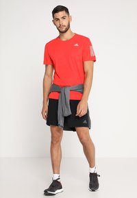 Camiseta deportiva roja con tres rayas blancas en el hombro izquierdo, combinada con pantalones cortos negros y un suéter gris atado a la cintura.