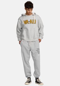 Felpa grigia con lettere a contrasto gialle e nere, tasca frontale. Pantaloni della tuta grigi con testo stampato e logo. Sneakers alte nere.