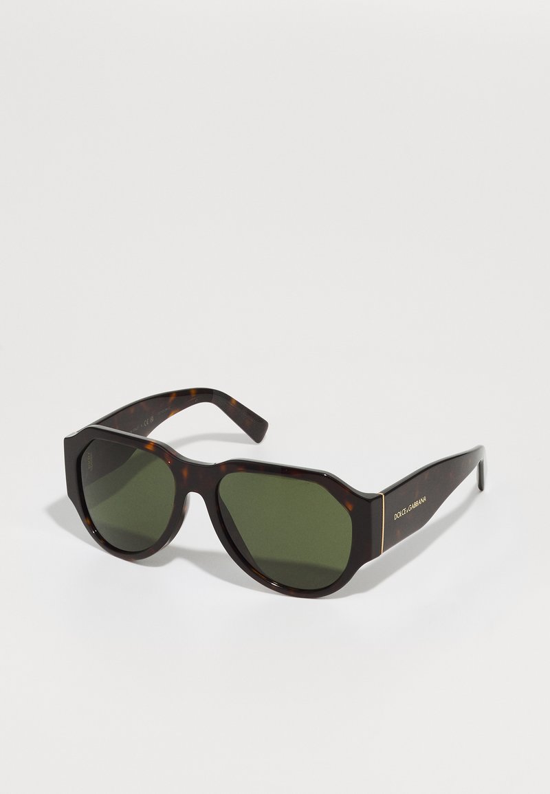 Sietu formas saulesbrilles ar zaļiem lēcas, lielizmēra astoņkantīgas formas, un zelta logo akcents uz tempļiem. Gluda plastmasas rāmja dizains.