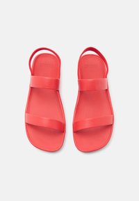 Κόκκινα σανδάλια με επίπεδη σόλα, με δύο φαρδιές λουριά κατά μήκος του ποδιού και ένα λουράκι στο πίσω μέρος για στήριξη. Κατασκευασμένα από λείο, ανθεκτικό υλικό.