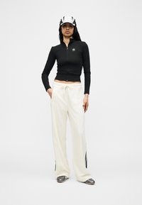 Γυναίκα που στέκεται με τα χέρια στις τσέπες, φοράει μαύρο μπουφάν με φερμουάρ, λευκό παντελόνι με ριγέ πλαϊνές ρίγες, ασημένια παπούτσια και μαύρη και άσπρη καπέλο.