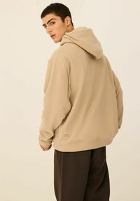 Beżowy polarowy hoodie z przednią kieszenią, luźnym fasonem i dużym kapturem. Posiada metkę z logo na dolnym lewym brzegu. W zestawie z brązowymi spodniami.