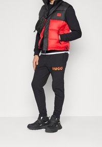 Homme portant une chemise noire à manches longues, une doudoune rouge et noire, un pantalon de survêtement noir avec un logo "HUGO" enflammé, des baskets noires et un sac bandoulière noir.