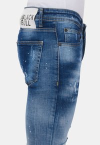 Verbleekte blauwe spijkerbroek met verweerde details, vijf zakken en een wit leren label met de tekst "BLACK BULL" op de tailleband.