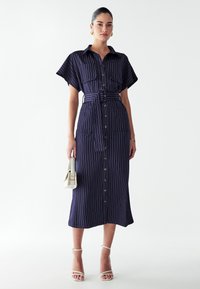 Pruunide triipudega nööpidega kleit lühikeste varrukatega, vöödiga taljes ja kahe esikotiga. Kanda strappy valgete sandaalide ja väikese kotti.