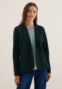 Vrouw in een donkere blazer over een lichtgrijze trui met blauwe jeans, staand tegen een effen beige achtergrond.