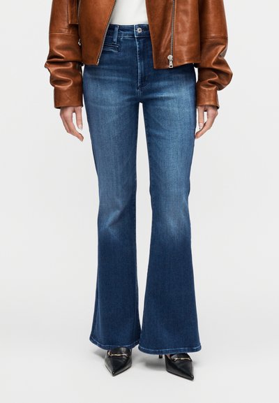 Μπλε τζιν παντελόνια καμπάνα με μέση μεσαίου ύψους, κατασκευασμένα από ύφασμα denim, με ελαφρύ ξεθώριασμα και καθαρές ραφές. Φοριούνται με μαύρες μυτερές γόβες.