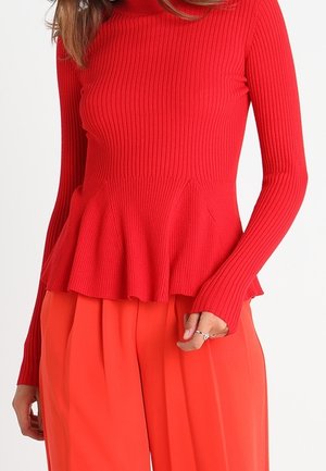 Frau trägt ein rotes, geripptes Langarmshirt mit Peplum-Saum und leuchtend orangefarbene, weit geschnittene Hosen, zeigt einen Teil ihres Oberkörpers und ihrer Arme.