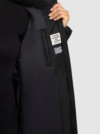 Musta täispuhutava jope siseosa, millel on tikitud mustriga kangas, sildiga, millel on kiri "Soe kallistus kangast", ning allosas logo.