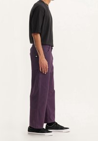 Baggy lila byxor i ett texturerat bomullstyg, med sidofickor och en liten logotyp; tillsammans med en svart t-shirt med kort ärm och svarta sneakers.