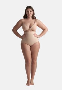Donna in piedi a piedi nudi con le mani sui fianchi, che indossa shapewear a vita alta color carne e un reggiseno abbinato, su uno sfondo neutro.