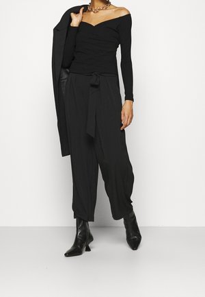 Donna con top nero scollato, pantaloni a gamba larga a vita alta con cintura, che tiene una giacca nera a spalla e stivaletti neri.