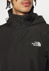 Nuori henkilö pukeutuneena mustaan North Face -takkiin, jossa on huppu, lähikuva takin kauluksesta ja rinnassa olevasta logosta.